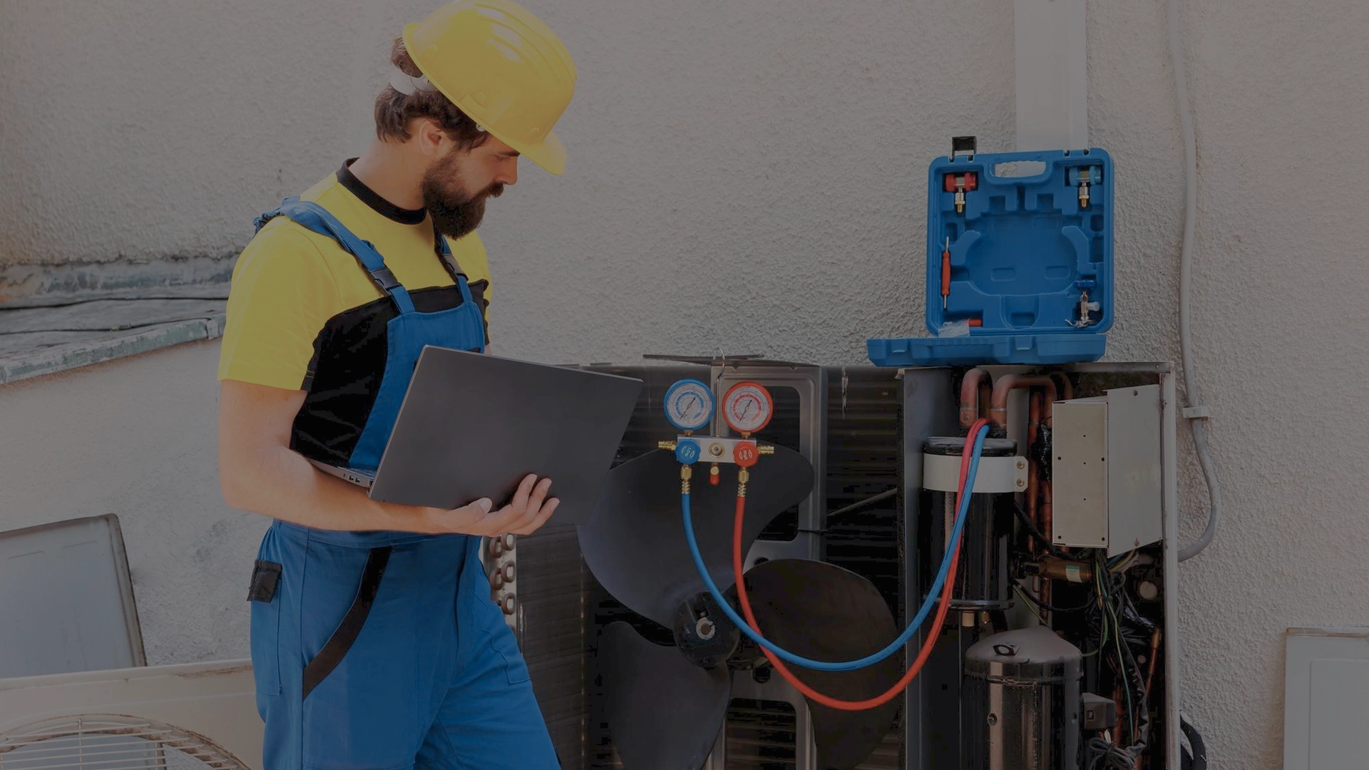 An HVAC technician in a yellow hard hat and blue uniform inspecting an open outdoor air conditioning unit with professional tools, showcasing expert HVAC maintenance services in Greater Boston, MA.