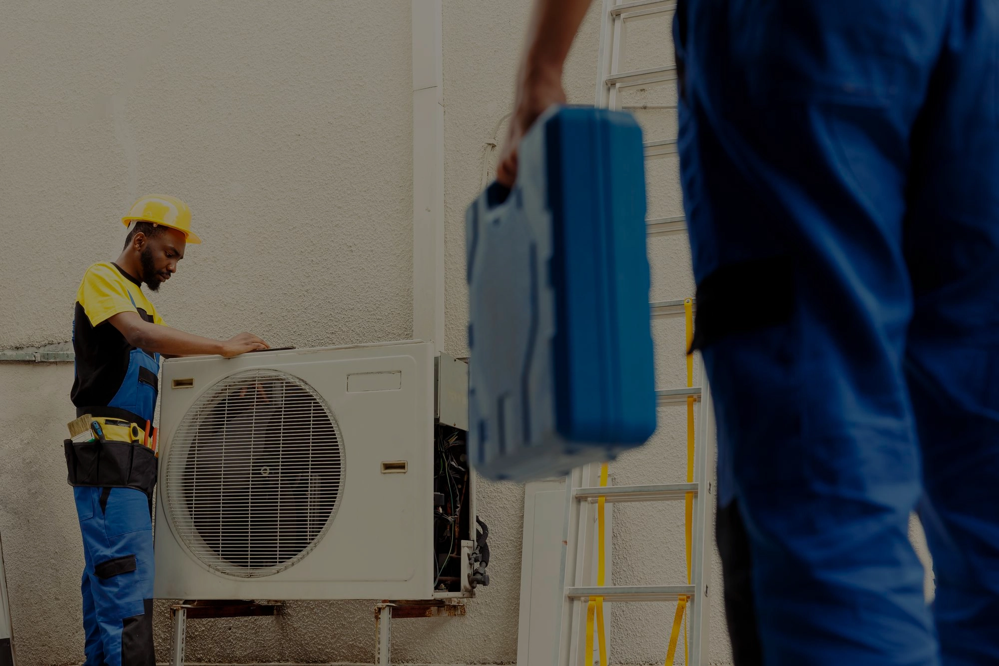 HVAC technician in a yellow hard hat inspecting an outdoor air conditioning unit while another technician carries a blue toolbox, highlighting teamwork in professional AC repair and maintenance services in Greater Boston, MA.