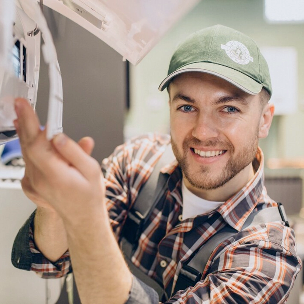 Smiling HVAC technician wearing a green cap and plaid shirt, performing maintenance on an indoor air conditioning unit to ensure reliable AC service and repair in Boston, MA.