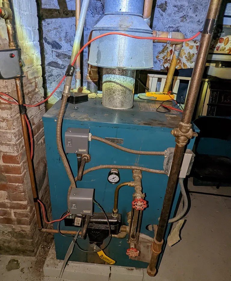 A detailed view of an older boiler system with a cylindrical metal vent, surrounded by copper and iron pipes. The setup includes manual valves, electrical components, and a pressure gauge.