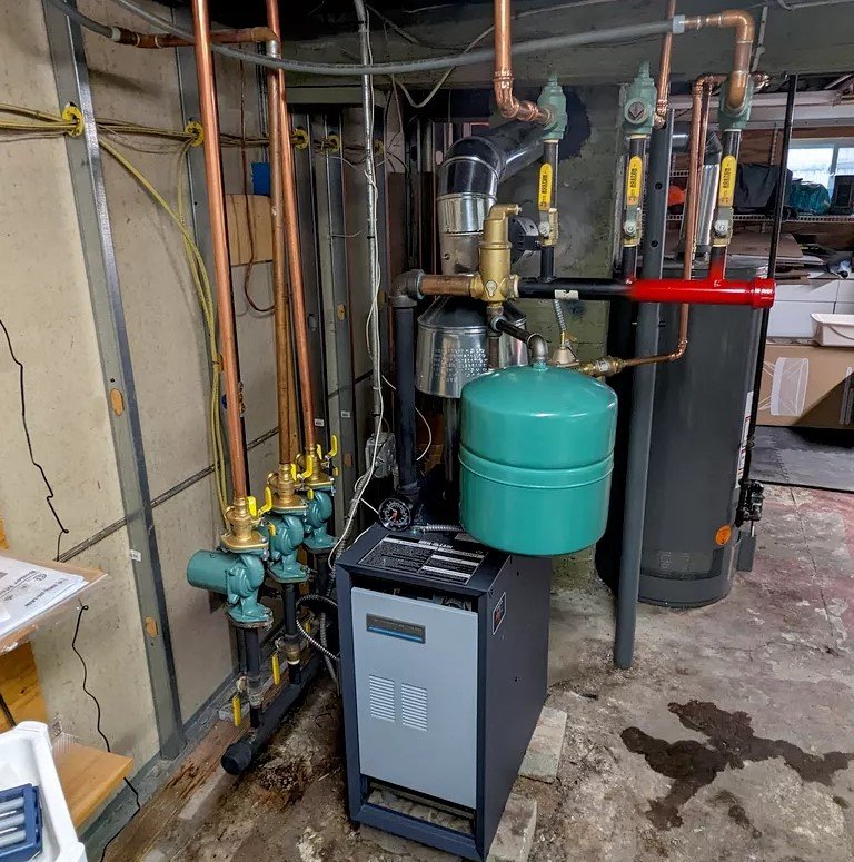 A basement utility area with a modern boiler installation. The system includes copper and iron pipes, a green expansion tank, and a tall silver exhaust vent, all neatly organized.